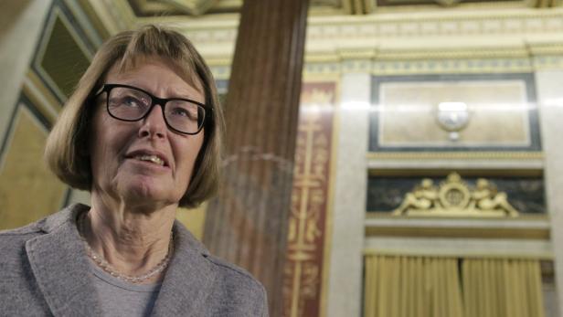 Eine Frau mit Brille und grauem Blazer steht in einem verzierten Raum.
