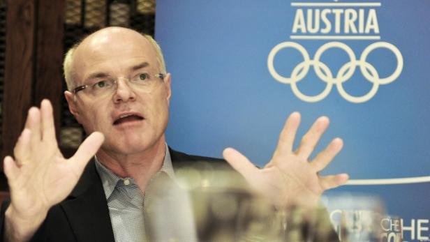 Ein Mann mit Brille gestikuliert vor einem blauen Hintergrund mit dem Schriftzug „Austria“ und den olympischen Ringen.