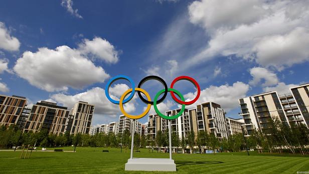 Die olympischen Ringe stehen auf einer Wiese vor modernen Wohngebäuden.