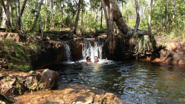 Zwei Personen schwimmen in einem natürlichen Pool unter einem kleinen Wasserfall.
