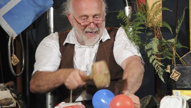 Ein Mann mit Brille schlägt mit einem Hammer auf einen roten und einen blauen Ballon.