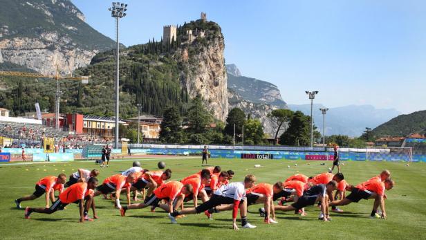 Eine Fußballmannschaft dehnt sich auf einem Rasenplatz vor einer Bergkulisse mit Burg.