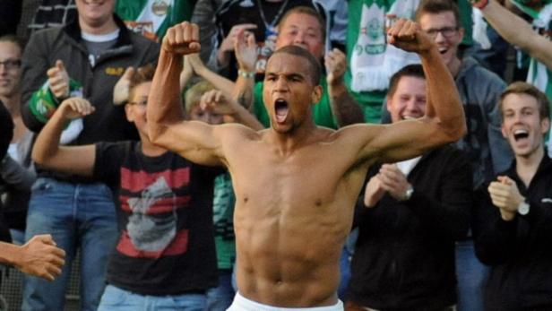 Ein jubelnder, muskulöser Mann steht vor einer feiernden Menschenmenge.