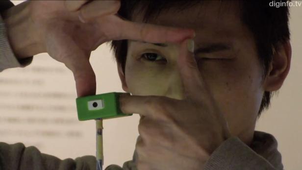 Eine Person blickt durch einen mit den Händen geformten Rahmen, an dem ein kleiner, grüner Gegenstand befestigt ist.