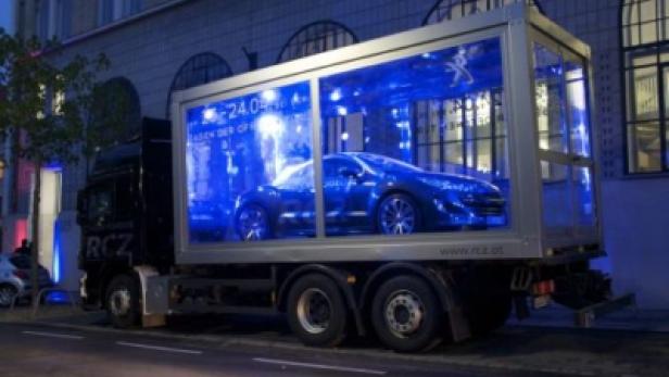 Ein Peugeot RCZ wird in einem gläsernen Ausstellungsraum auf einem LKW transportiert.