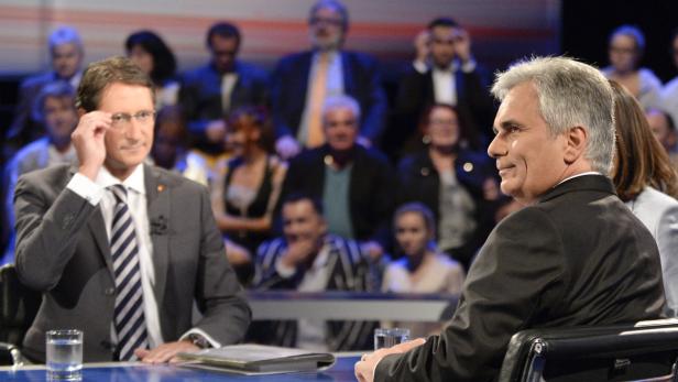Zwei Männer in Anzügen sitzen vor einem Publikum auf einer Bühne.