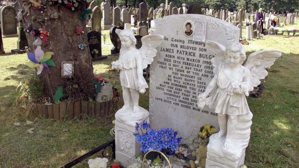 Auf einem Friedhof befindet sich der Grabstein von James Patrick Bulger, geschmückt mit Blumen und Engelsstatuen.