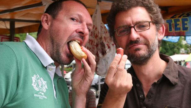Zwei Männer essen Würstchen an einem Imbissstand.