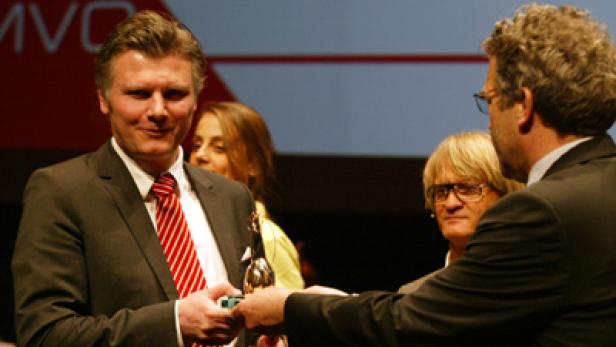 Ein Mann im Anzug überreicht einem anderen Mann eine goldene Trophäe.