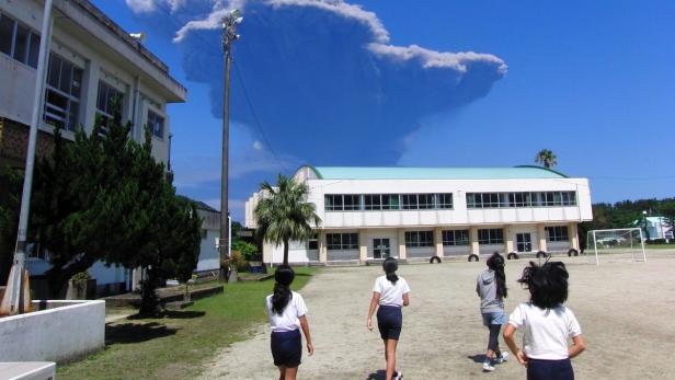 Schülerinnen gehen auf einem Schulhof, im Hintergrund ein Gebäude und eine große Rauchwolke.