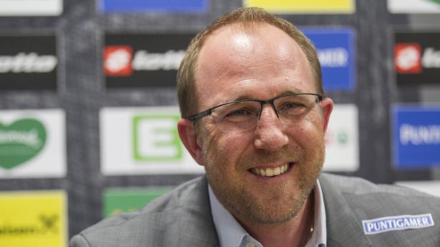 Ein lächelnder Mann mit Brille und Sakko vor einer Wand mit Logos.