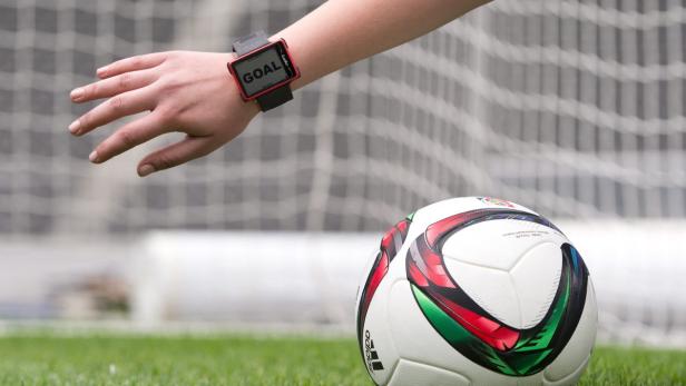 Eine Hand mit einer Smartwatch, die „GOAL“ anzeigt, neben einem Fußball auf einem Rasenplatz.