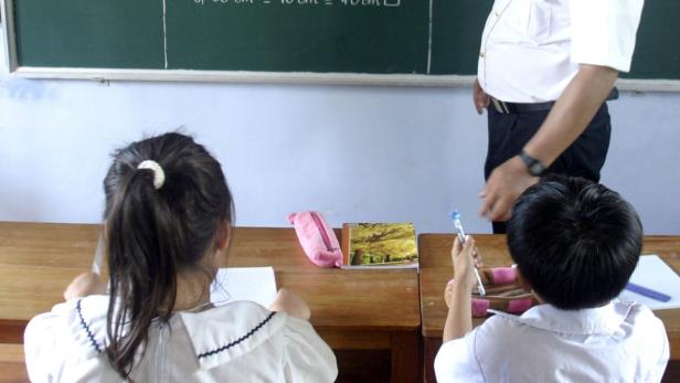Zwei Schulkinder sitzen in einem Klassenzimmer, während ein Lehrer an der Tafel steht.
