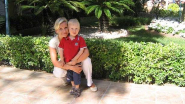 Eine Frau umarmt einen kleinen Jungen vor einer grünen Hecke und Palmen.
