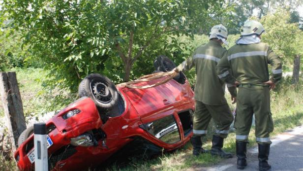 Ein rotes Auto liegt nach einem Unfall auf der Seite, während zwei Feuerwehrleute die Situation begutachten.