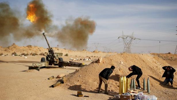Eine Haubitze feuert in einer Wüstenlandschaft, während Männer in der Nähe Munition handhaben.