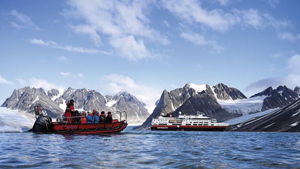 Ein rotes Schlauchboot mit Touristen fährt vor einer schneebedeckten Berglandschaft und einem Kreuzfahrtschiff.