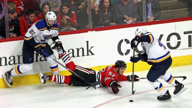 Ein Eishockeyspieler der Chicago Blackhawks liegt auf dem Eis, während zwei Spieler der St. Louis Blues sich nähern.