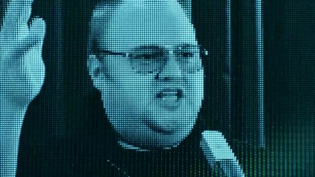 Ein Mann mit Brille spricht in ein Mikrofon vor einem blaugrünen Bildschirm.