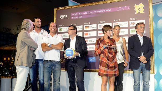 Personen stehen vor einer Tafel mit dem Spielplan für das olympische Beachvolleyballturnier der Männer.