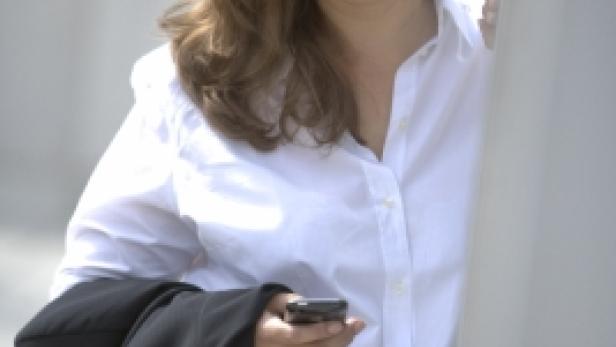 Eine lächelnde Frau in weißem Hemd hält ein Handy und einen Blazer in der Hand.