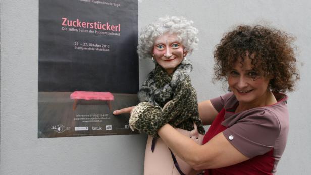 Eine Frau mit lockigen Haaren hält eine Puppe vor einem Plakat für „Zuckerstückerl“.