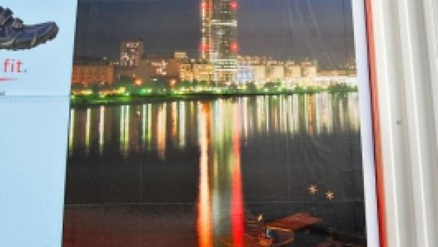 Ein Plakat zeigt ein nächtliches Stadtbild von Wien mit dem DC Tower, der sich im Wasser spiegelt.