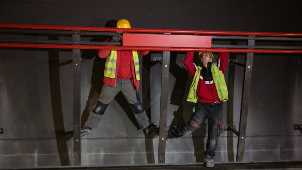 Zwei Bauarbeiter mit Schutzhelmen montieren eine rote Konstruktion an einer Wand.