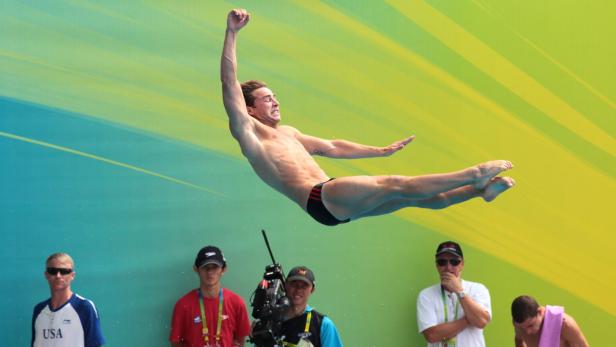 Ein männlicher Turmspringer in der Luft vor einem gelb-türkisfarbenen Hintergrund.