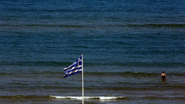 Eine griechische Flagge weht am Strand, während eine Person im Meer schwimmt.