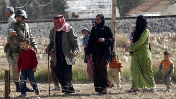 Eine Gruppe von Menschen, darunter Kinder und ein Soldat, geht an einem Stacheldrahtzaun entlang.