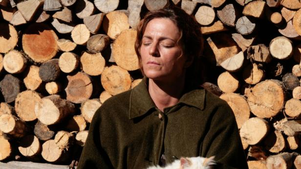 Eine Frau mit geschlossenen Augen lehnt an einem Holzstapel, eine Katze im Arm.