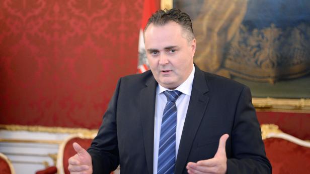 Ein Mann im Anzug gestikuliert vor einem roten Hintergrund.