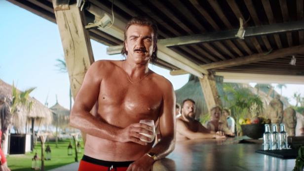 Ein Mann mit Schnurrbart steht barfuß an einer Bar im Freien und hält ein Glas Milch.