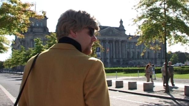 Ein Mann mit Sonnenbrille und Jackett vor dem Reichstagsgebäude in Berlin.
