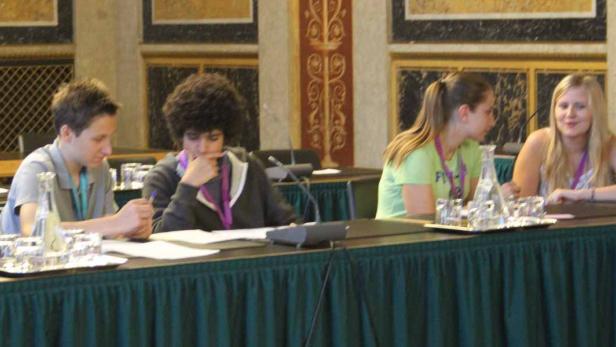 Vier junge Leute sitzen an einem Tisch mit Mikrofonen und Wasserflaschen.