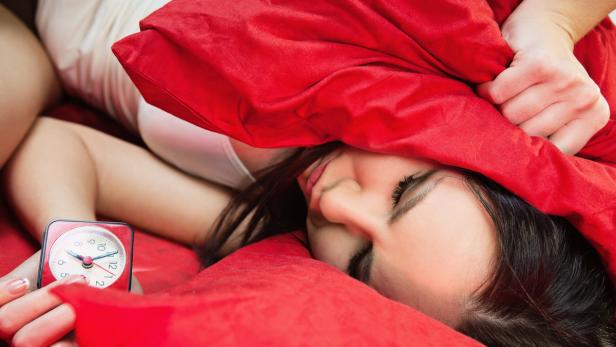 Eine Frau schläft mit einem roten Kissen über dem Kopf, während ein Wecker klingelt.