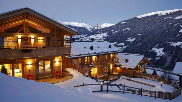 Mehrere beleuchtete Holzhäuser stehen im Schnee vor einer Bergkulisse.