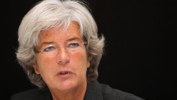 Eine Frau mit grauen Haaren und Brille spricht vor einem dunklen Hintergrund.