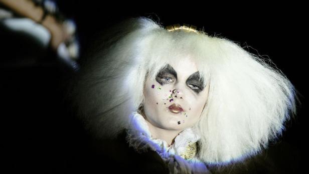 Eine Person mit weißer Perücke und auffälligem Make-up blickt in die Kamera.