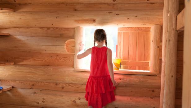 Ein Mädchen im roten Kleid schaut aus einem Fenster in einem Holzhaus.