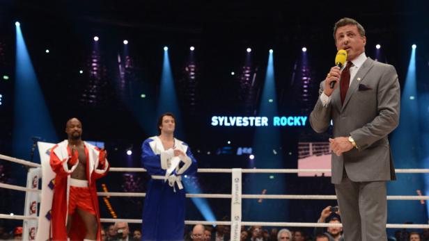 Sylvester Stallone spricht im Boxring, während Boxer in Bademänteln im Hintergrund stehen.