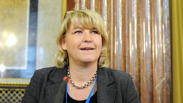 Eine blonde Frau mit einer silbernen Halskette spricht in ein Mikrofon.