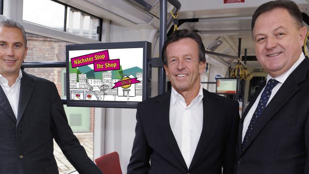 Drei Männer in Anzügen stehen in einer Straßenbahn neben einem Bildschirm mit der Aufschrift „Nächster Stop Ihr Shop“.