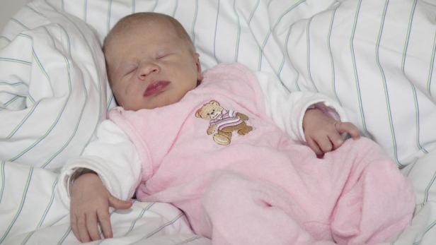 Ein neugeborenes Baby schläft in einem rosa Strampler mit Teddybär-Motiv auf einem Bett.