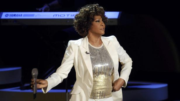 Whitney Houston tritt in einem weißen Anzug mit Mikrofon in der Hand auf einer Bühne auf.
