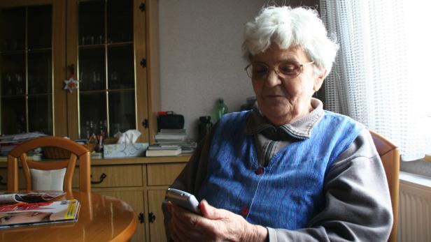 Eine ältere Frau sitzt am Tisch und benutzt ein Mobiltelefon.