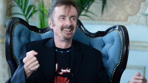 Ein lächelnder Mann mit Bart sitzt auf einem verzierten blauen Sessel.