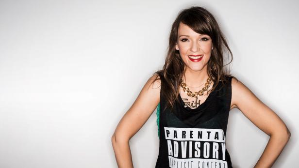 Eine lächelnde Frau mit rotem Lippenstift und einem „Parental Advisory“-Top posiert vor einer weißen Wand.
