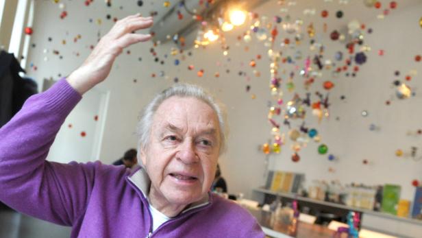 Ein älterer Mann mit erhobenem Arm steht vor einer Wand mit bunten Dekorationen.